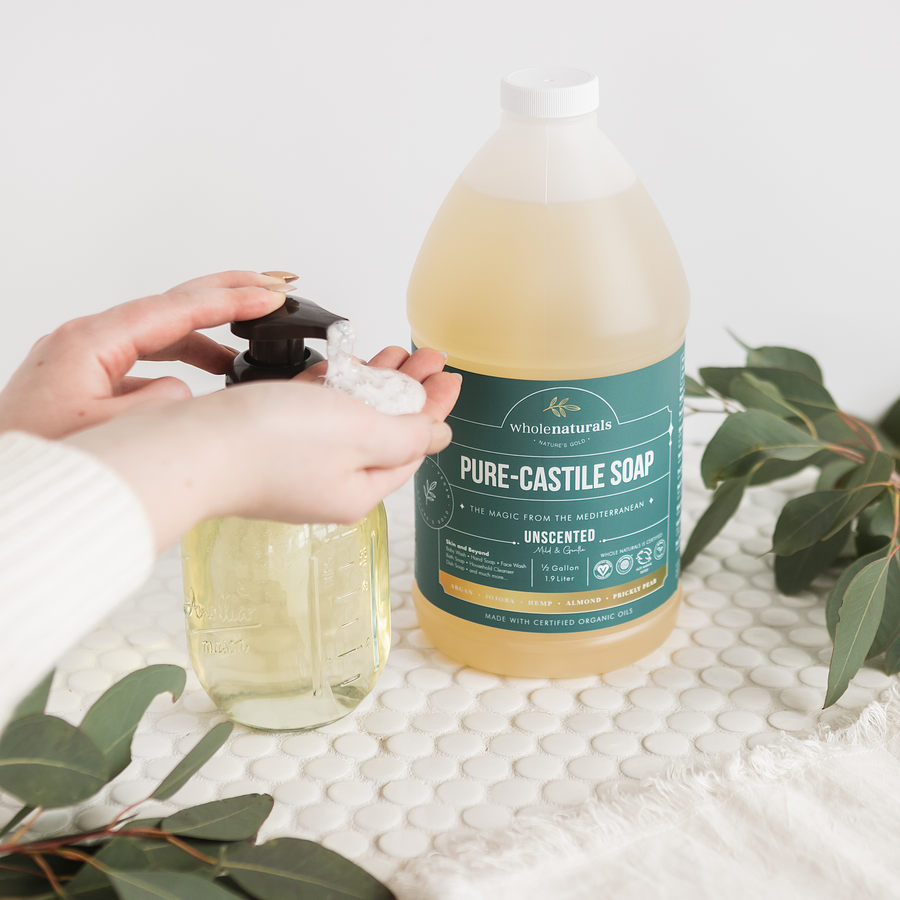 Hands pumping Pure-Castile Soap into a glass dispenser beside the 64 oz bottle, with eucalyptus on a tiled counter