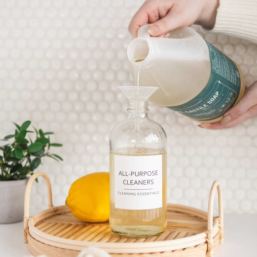 Pouring Pure-Castile Soap through a funnel into a glass bottle labeled All-Purpose Cleaners, with a lemon and a plant on a rattan tray